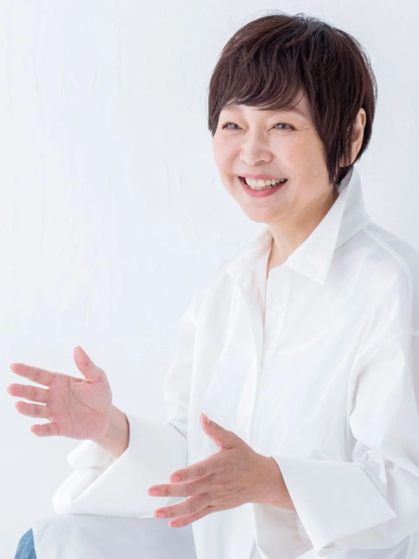 白いシャツを着たショートヘアの日本人女性が、笑顔で両手を広げて話しているようなポーズをとっているポートレートです。