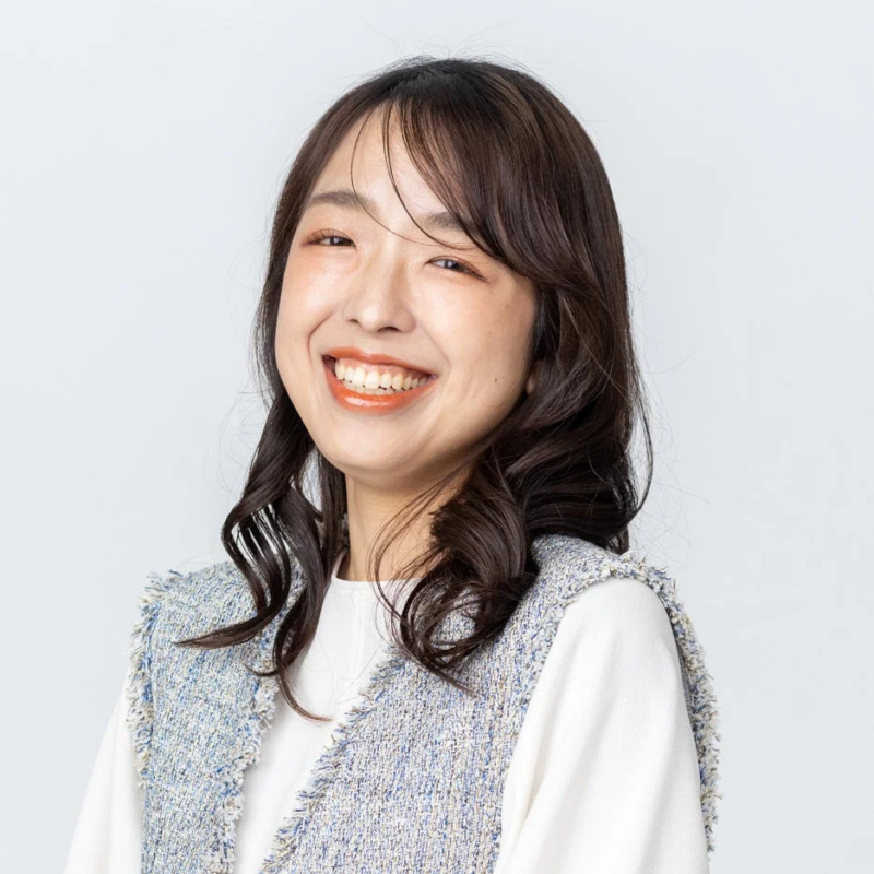 白い背景の前で、カメラに向かって満面の笑みを浮かべる若いアジア人女性のポートレートです。長いダークヘアで、白いトップスの上に青と白のツイード風ベストを着用しており、明るく親しみやすい印象を与えます。