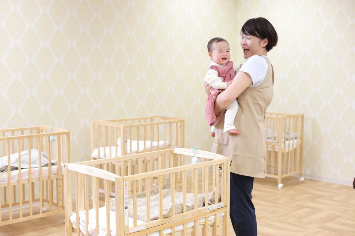 保育士らしき女性が笑顔の赤ちゃんを抱っこしており、複数のベビーベッドが置かれた保育施設の一室の様子