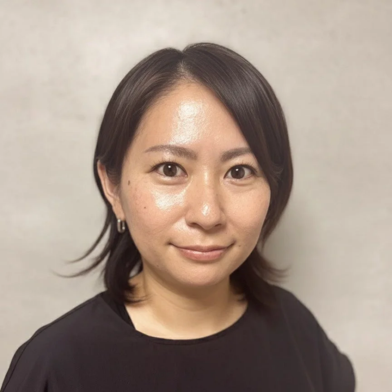 カメラに視線を向けるアジア人女性のポートレート。肩までの長さの黒髪で、黒い服を着用し、穏やかな笑顔を浮かべている。背景はシンプルな明るい色調の壁です。