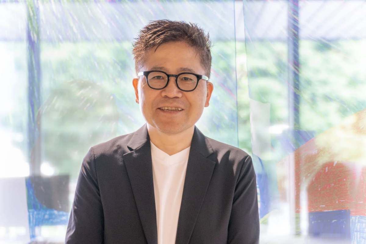 眼鏡をかけた男性が、黒いジャケットと白いTシャツを着用し、カメラに向かって笑顔を見せているポートレート写真