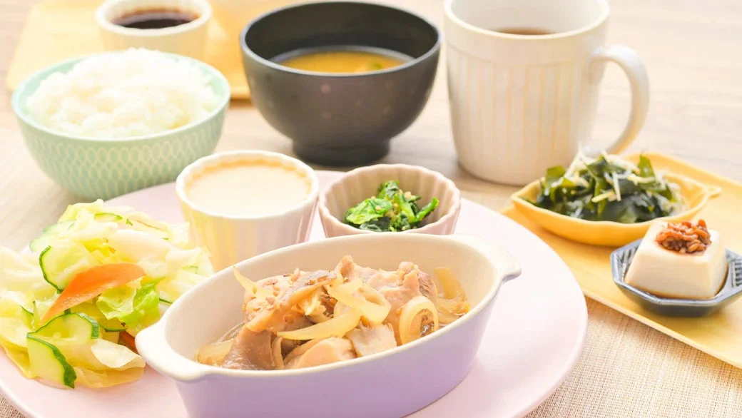 ご飯、味噌汁、主菜の鶏肉と玉ねぎの煮物、キャベツとキュウリのサラダ、ほうれん草のおひたし、わかめの和え物、冷奴などが並んだ、バランスの取れた和食の定食
