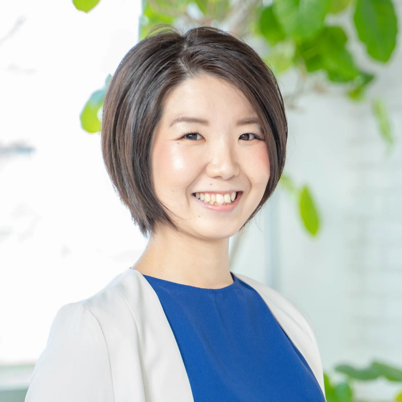 ショートヘアの女性が明るい笑顔でカメラを見ているポートレート写真です。青いトップスに白いジャケットを羽織っており、背景にはぼかされた緑の植物が見えます。親しみやすい印象を与えます。
