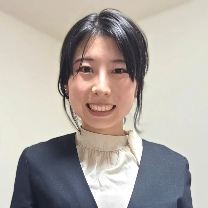 笑顔でカメラを見つめる若い女性のポートレート。黒いジャケットとベージュのブラウスを着用しており、清潔感のある印象です。白い背景で撮影されています。