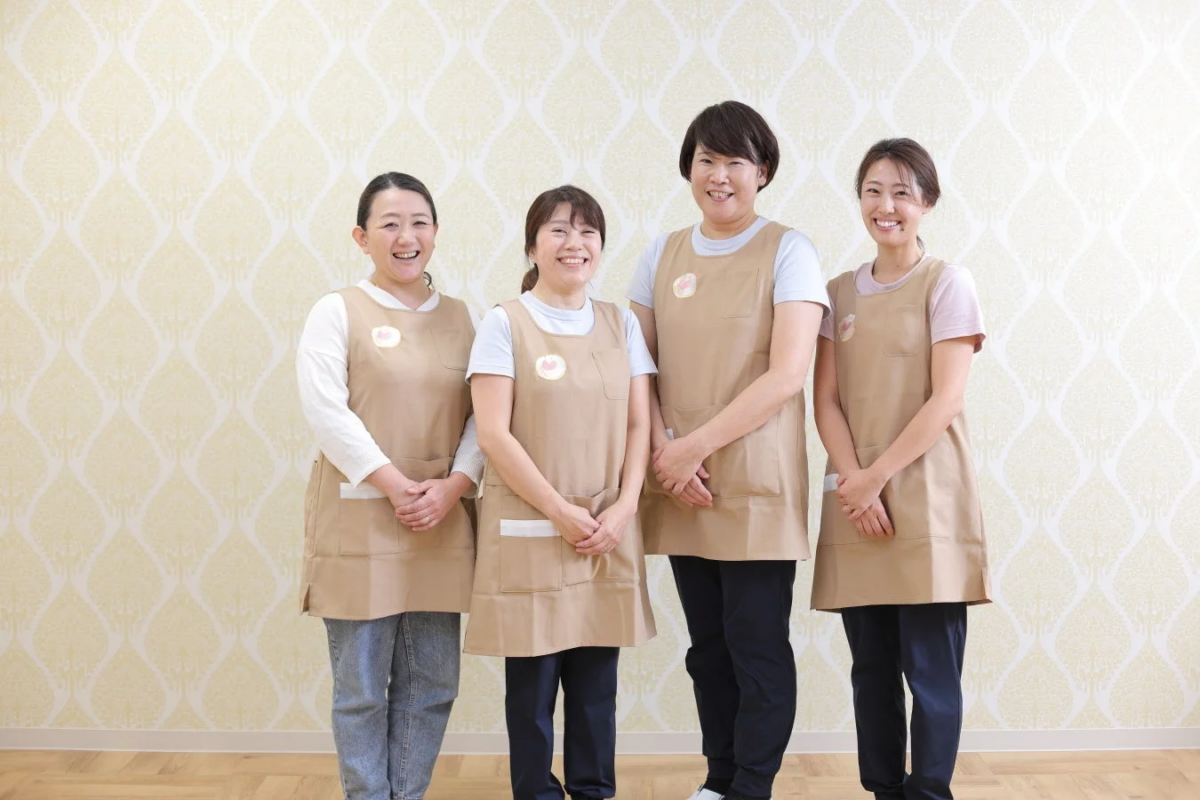 ベージュのエプロンを着た4人の日本人女性が、笑顔でカメラに向かって並んでいます
