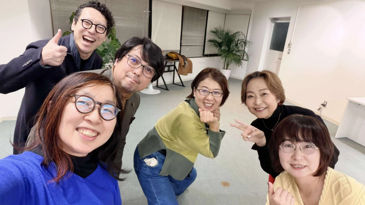 オフィスのような明るい室内で、男女6人が集まって笑顔でセルフィーを撮っている集合写真です。