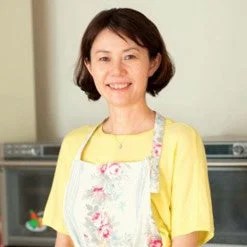 黄色のTシャツと花柄のエプロンを着用し、笑顔でカメラを見つめるアジア人女性のポートレートです。背景にはキッチンらしき室内が写っています。