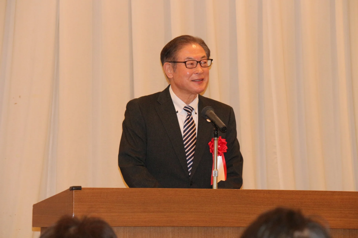 スーツを着た男性が、マイクのある演台でスピーチをしている様子です。背景はシンプルな白いカーテンで、フォーマルなイベントや会議の雰囲気を示しています。男性は笑顔で話しています。