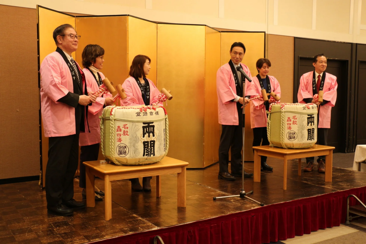 金屏風を背景にしたステージで、ピンクのはっぴを着た男女数人が鏡開きを行っている様子です。日本酒の酒樽を前に木槌を持ち、祝賀ムードが漂うイベント風景が写し出されています。