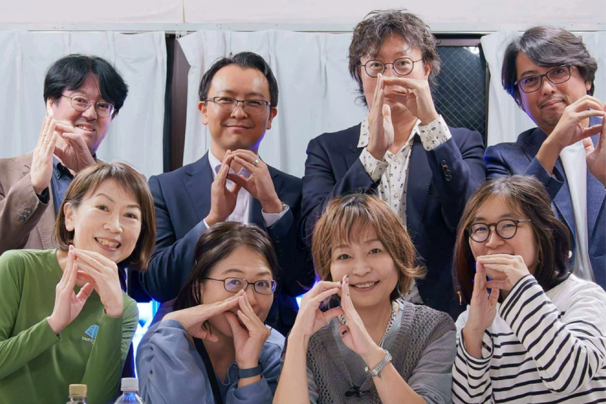 笑顔の男女8人が顔の前で手サインをしてポーズをとる集合写真です。