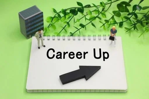 ノートに「Career Up」という文字と上向きの矢印が描かれ、その周りにビジネスマンのミニチュア人形が配置されています。キャリアアップや目標達成への意欲を表すイメージです。