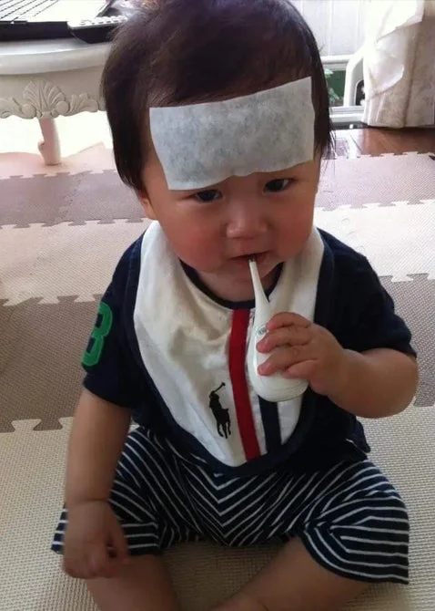 おでこに熱さましシートを貼り、体温計を口にくわえている赤ちゃんの写真