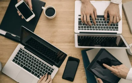 複数の人がノートパソコン、スマートフォン、タブレットといったデジタルデバイスを使い、木製のデスクで共同作業や会議をしている様子を上から捉えた画像です。ビジネスシーンや集中して作業する風景が伺えます。