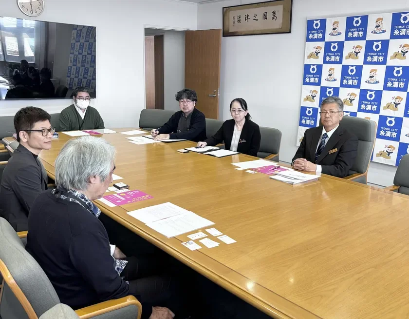 糸満市の会議室で、数名が大きな木製のテーブルを囲んで会議を行っている様子。壁には「萬國津梁之」の額と、糸満市のロゴが入った背景が確認できる。