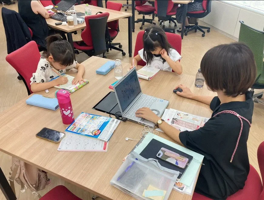 教室のような空間で、2人の幼い女の子と1人の大人の女性がテーブルを囲んで座っている