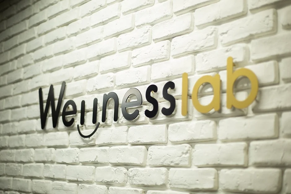 白いレンガの壁に「Wellnesslab」と書かれた立体的なロゴが取り付けられている画像です。Wellnessの部分は黒色で、labの部分は黄色の文字で表現されています。