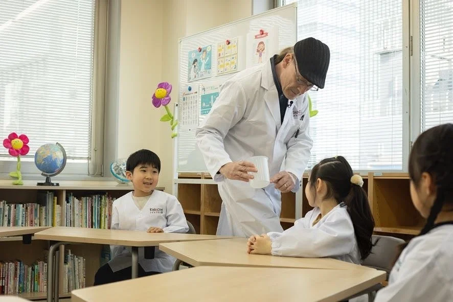 白衣を着た先生が、同じく白衣を着た小学生の子供たちに科学実験を教えている教室の光景です。子供たちは興味津々な様子で実験を見つめています。