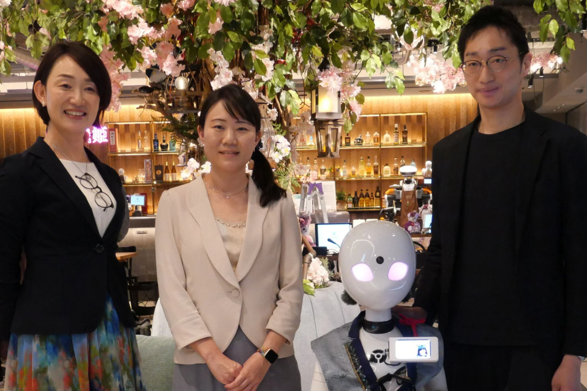 ピンクの花で飾られた空間で、2人の女性、1人の男性、そして白い頭部のロボットが並んで立っています。背景にはボトルが並んだ棚が見えます。