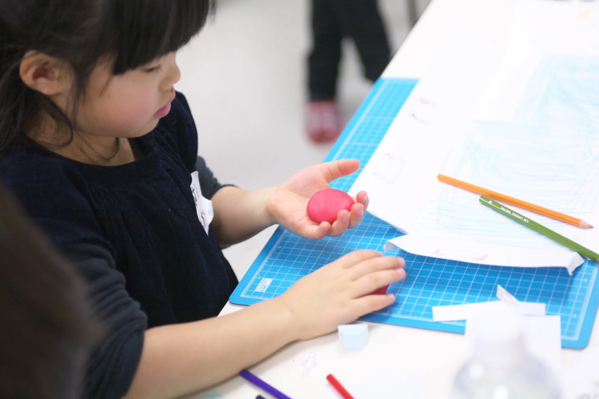 幼い女の子がテーブルでクラフト活動に集中しています。ピンク色の粘土を手に持ち、青いカッティングマットの上で紙や色鉛筆を使って作業しています。