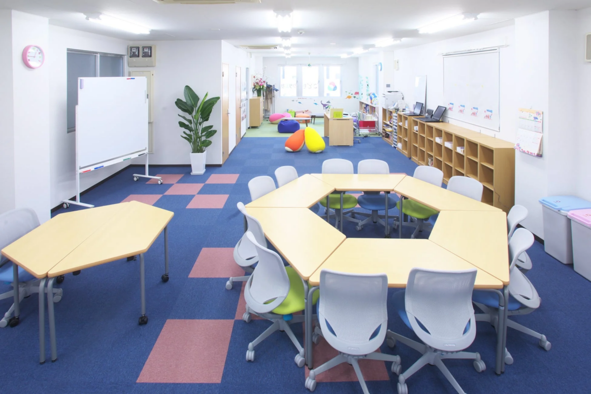 明るくモダンなデザインの多目的ルームで、学習や会議、共同作業に適しています。六角形のテーブルと椅子が中央に配置され、奥にはカラフルなクッションがあるリラックススペースも見えます。青いカーペットとピンクの市松模様が特徴的です。