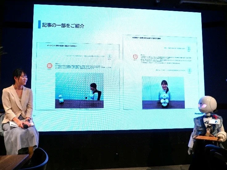 プレゼンテーション会場で、女性がマイクを持ち、大型スクリーンには記事の一部が紹介されています。スクリーンの横には人型ロボットが立っており、イベントや新しいユーザー体験に関する講演が行われている様子です。