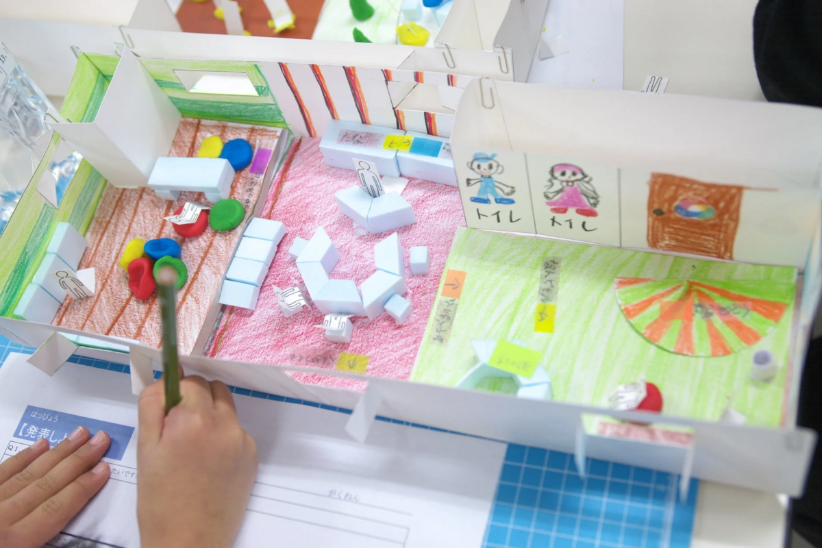子供が作成した部屋のジオラマが写っており、手前に「発表しよう」と書かれたプリントと鉛筆を持つ手が映っています。ジオラマにはミニチュアの家具や粘土、手描きのイラストがあり、「トイレ」「たな」「へやづくりゲーム」などの文字が見えます。これは学校の学習活動で、創造的な作品作りと発表の準備をしている様子を示しています。