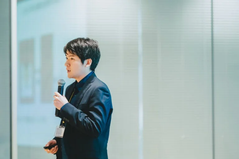 スーツを着用した男性がマイクとプレゼンターを持ち、真剣な表情で話しています。ビジネスイベントや会議での講演風景と思われます。