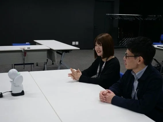 オフィスのような空間で、男女2人が白いテーブルを囲み、小型の白いロボットを前に会話している様子です。女性は笑顔で身振り手振りを交えており、活発な議論が行われているようです。