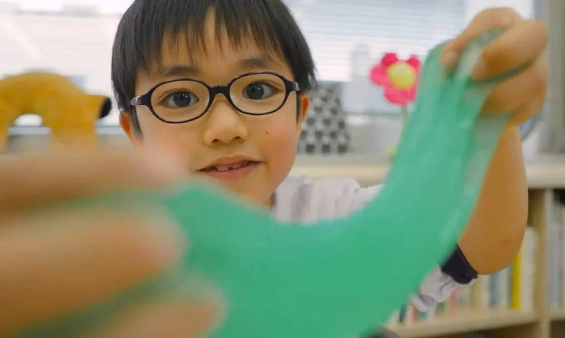 眼鏡をかけた男の子が、緑色のスライムを両手で伸ばしながらカメラを見ています。楽しそうな笑顔で、室内で遊んでいる様子が伝わります。