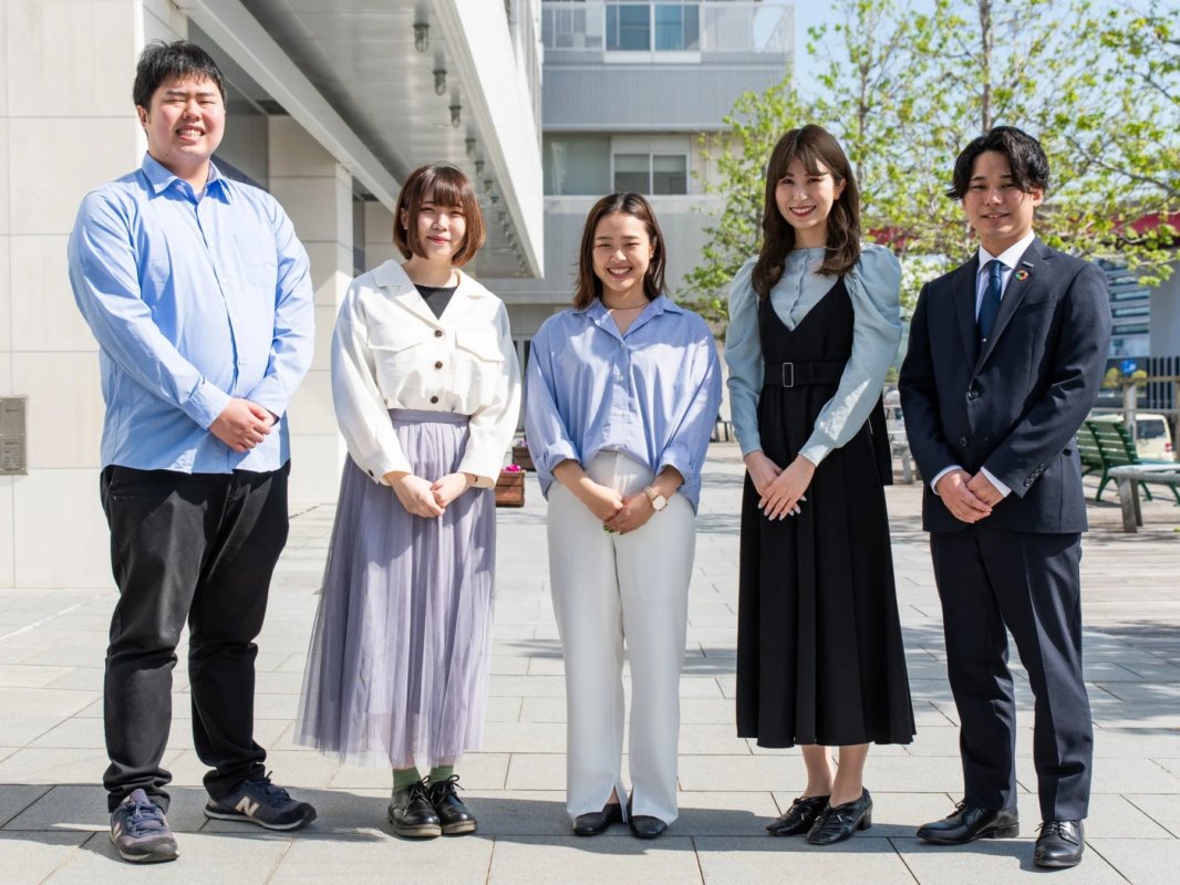 屋外で笑顔を見せる5人の若い日本人男女の集合写真です。カジュアルな服装とスーツ姿のメンバーが混在し、和やかな雰囲気が伝わります。