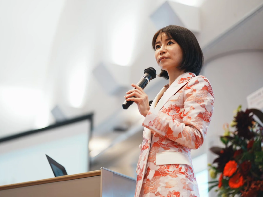 花柄のジャケットを着た女性がマイクを持ち、壇上で講演を行っている様子