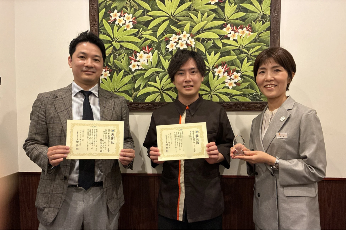 表彰状とトロフィーを手に記念撮影する3人のビジネスパーソン