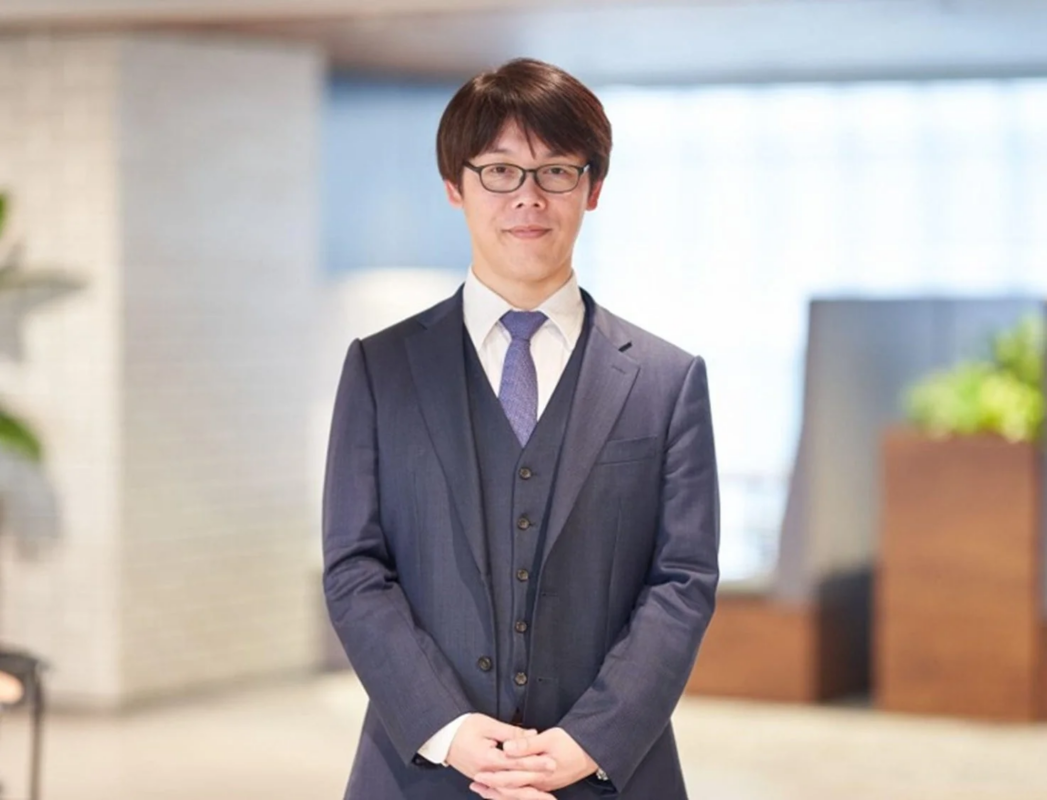 紺色のスーツと眼鏡をかけた男性が、室内で正面を向いて立っている画像