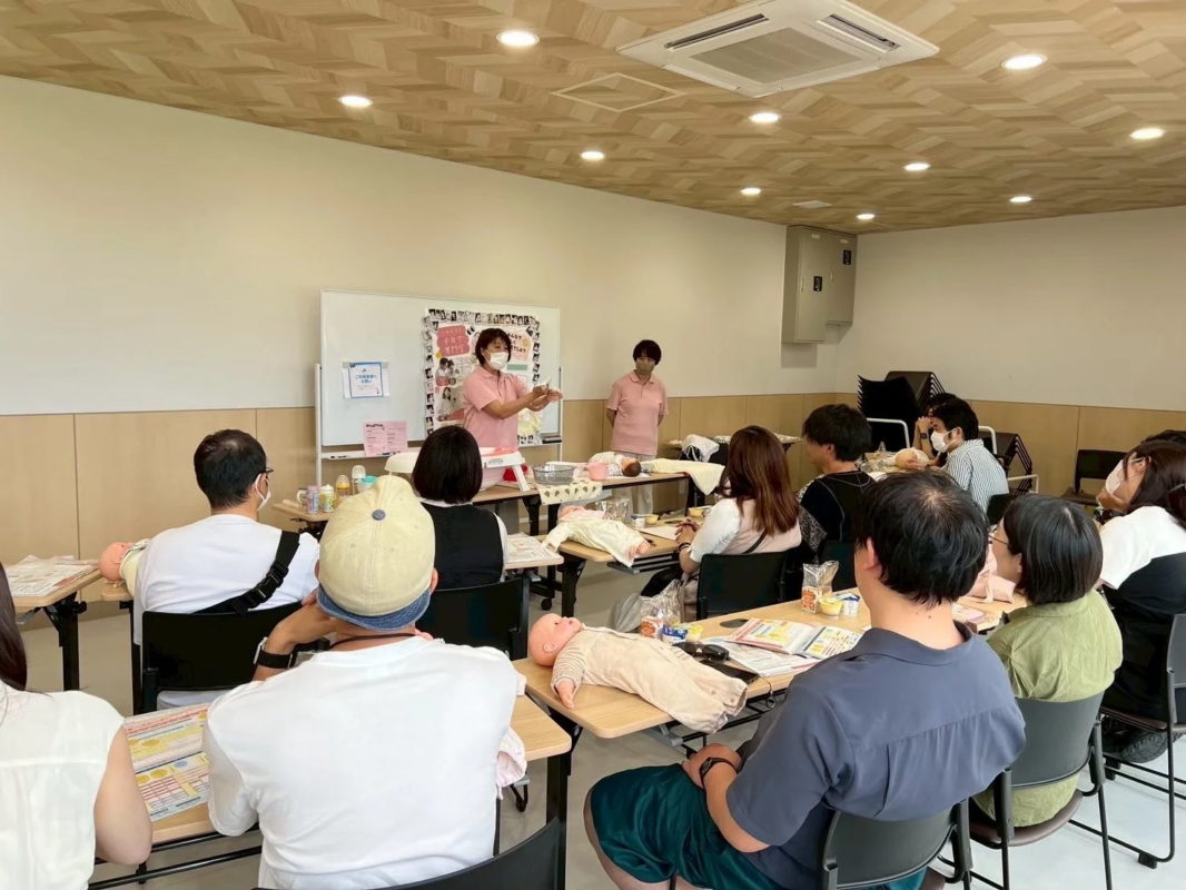 室内で行われている育児教室の様子です。講師が前に立ち、参加者たちは机に置かれた赤ちゃんの人形を使って、育児に関する指導を受けています。新生児ケアについて学んでいるようです。