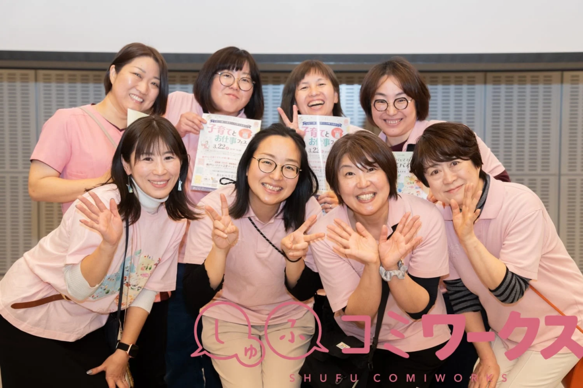 ピンクのTシャツを着た8人の女性たちが笑顔でポーズをとっている集合写真です。数人が「子育てと お仕事フェス」と書かれたチラシを持っており、和気あいあいとした雰囲気が伝わります。
