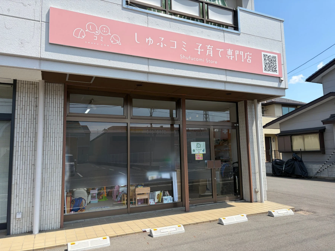 ピンクの看板が特徴的な「しゅふコミ 子育て専門店」の店舗外観です。大きなガラス窓からは店内の様子がうかがえ、子ども向けのアイテムが見えます。看板には英語表記とQRコードがあり、窓には「すべての子どもに笑顔を」「110番の家」と書かれたポスターが貼られています。